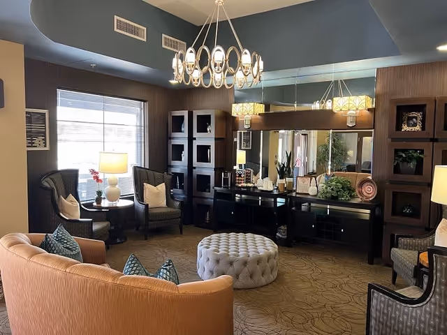 A cozy living room area with a curved beige sofa adorned with blue patterned pillows, two dark armchairs with light cushions, a round tufted ottoman in the center, and a side table with a lamp and a small plant near a window. The room features a modern chandelier, a large mirror above a black console table decorated with plants and decorative items, and dark wooden shelving units on either side.