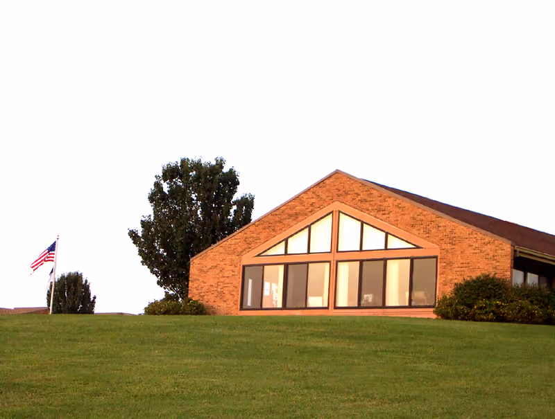 Brick building with large triangular and rectangular windows on a grassy hill beside an American flag.