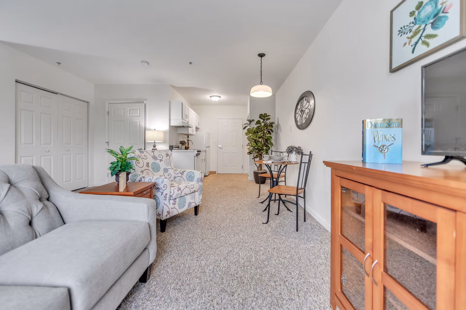 A bright and clean living area in a senior living facility featuring a light gray sofa, a patterned armchair, a small wooden side table with a plant, a round dining table with two chairs, a large wall clock, a wooden TV stand with a television and a book, and a small kitchen area in the background with white cabinets and appliances.