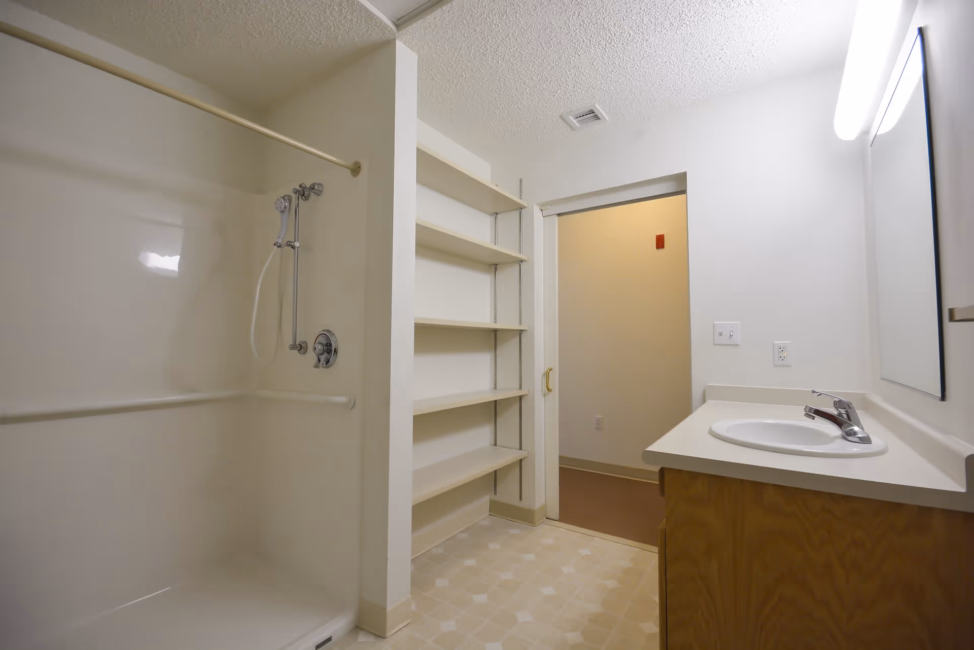 A clean bathroom with a shower area featuring a handheld showerhead and grab bars, open shelving next to the shower, a sink with a countertop and a mirror above it, and a doorway leading to another room.