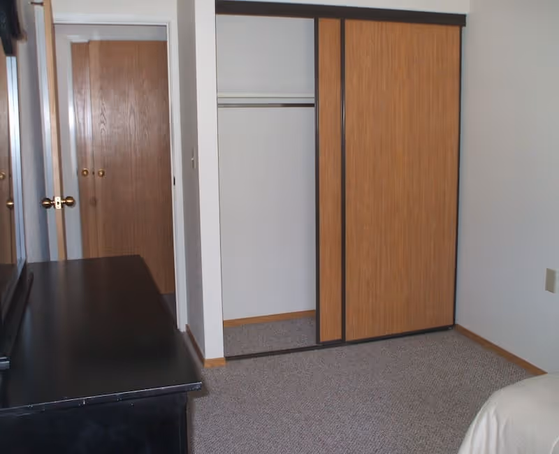 A bedroom corner featuring a partially open wooden sliding closet door revealing an empty closet. Next to the closet is a closed wooden door with brass handles. A black dresser with a mirror is visible on the left side, and part of a bed with a white bedspread is seen on the right. The room has beige carpet and white walls with wooden baseboards.