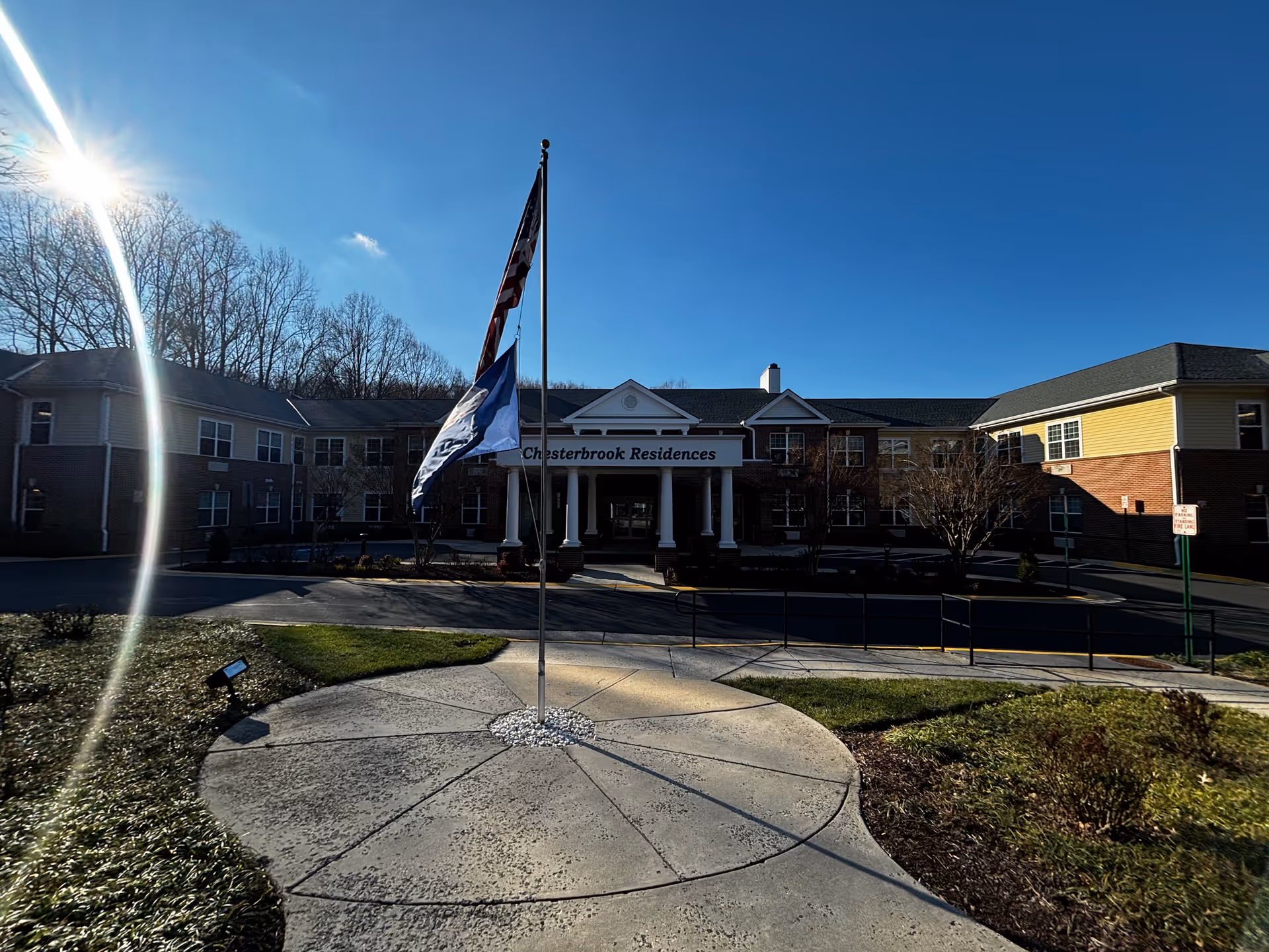 Exterior front view of Chesterbrook Residences building with a circular driveway and two flagpoles displaying the American flag and another flag. The building is two stories with a brick and siding facade under a clear blue sky.