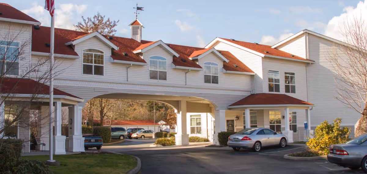 Exterior view of a white two-story building with a red roof, featuring an archway driveway and several parked cars in front. There is a flagpole with an American flag on the left side and some trees and bushes around the building.