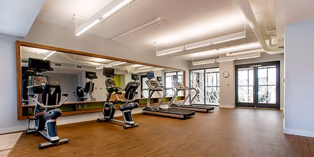 A fitness room with exercise equipment including stationary bikes and treadmills. The room has wooden flooring, large mirrors on one wall, and windows with natural light coming in. The ceiling has exposed pipes and fluorescent lighting.
