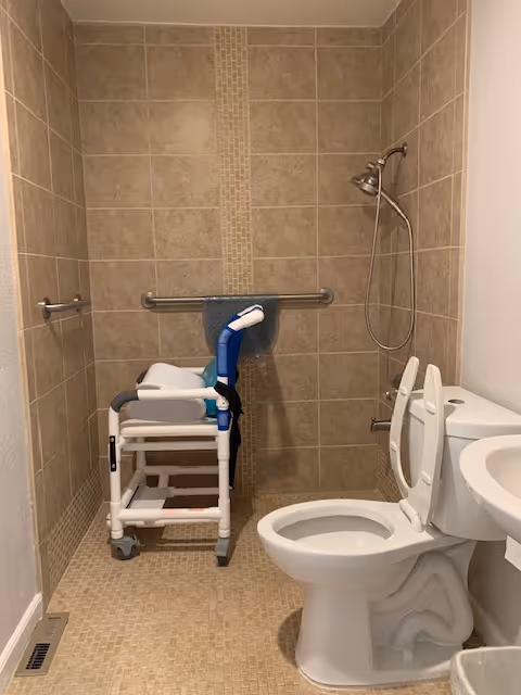A small bathroom with beige tiled walls and floor, featuring a white toilet with the lid open, a white sink, a shower area with a silver showerhead, and a white shower chair with blue handles. There is a grab bar mounted on the wall behind the shower chair with a blue towel hanging on it.