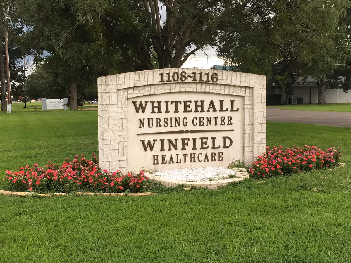 A stone sign on a grassy area with pink flowers planted around its base. The sign reads '1108-1116 WHITEHALL NURSING CENTER WINFIELD HEALTHCARE'. Trees and a road are visible in the background.