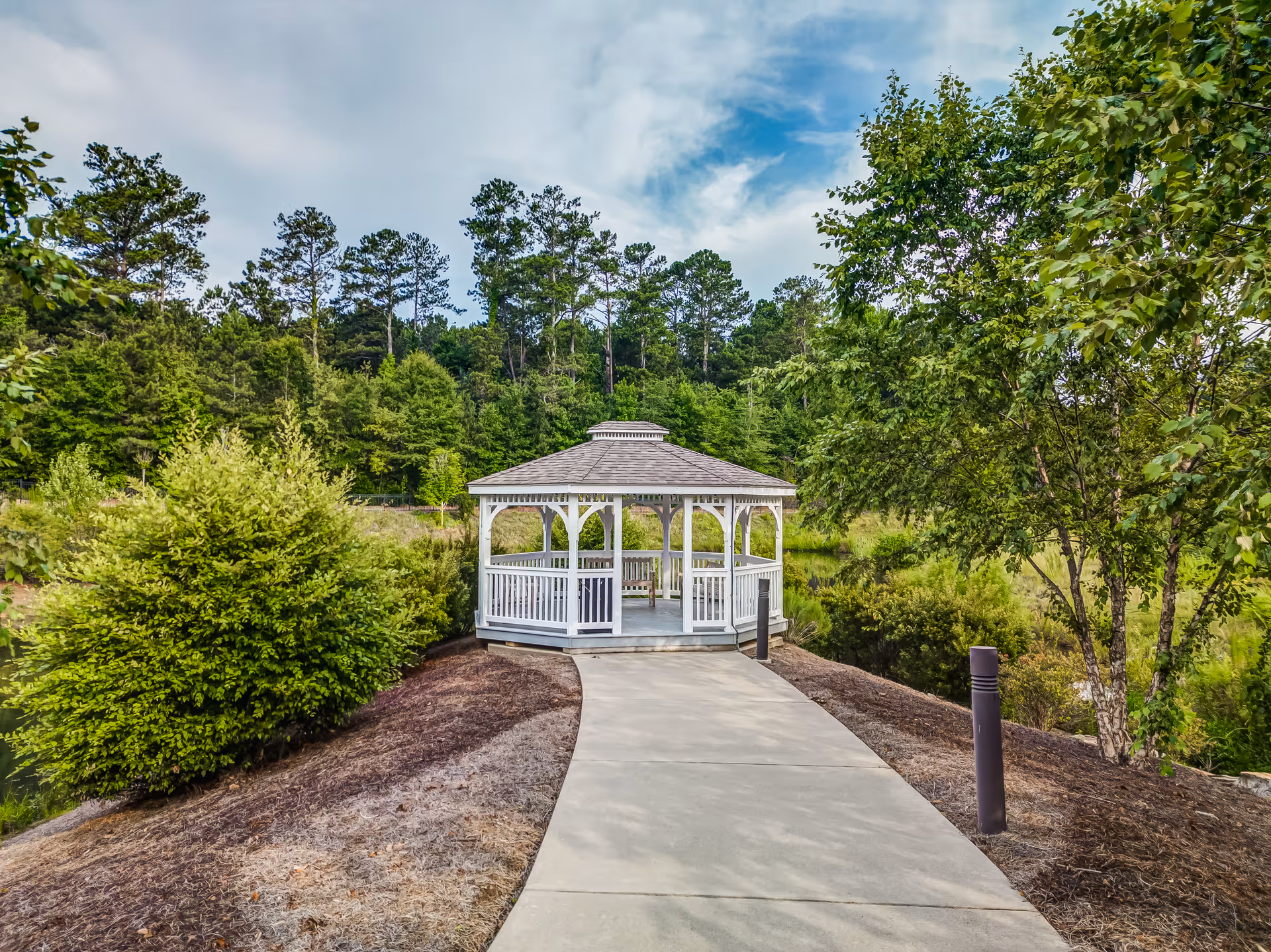 A paved walkway leading to a white wooden gazebo surrounded by green trees and bushes under a partly cloudy sky.