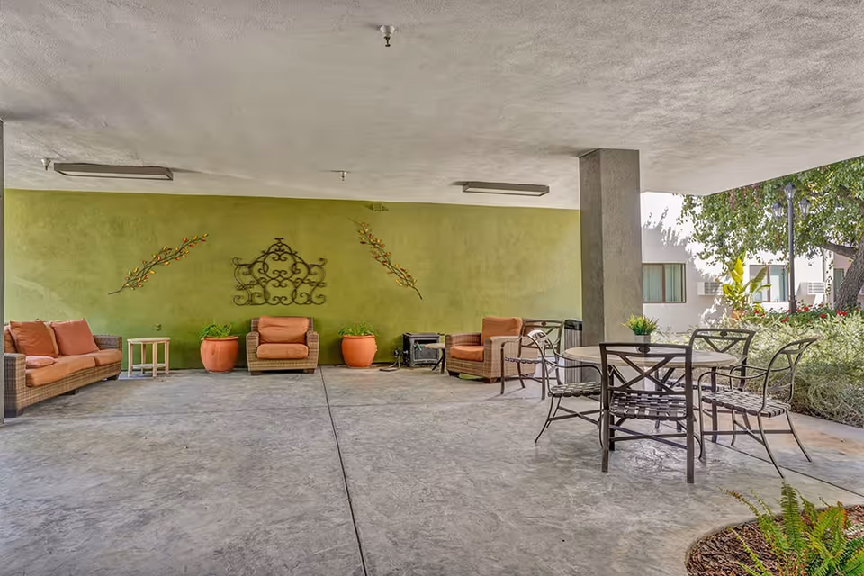 Covered outdoor seating area with cushioned wicker chairs and a sofa arranged against a green wall decorated with metal wall art and potted plants. A round metal table with four chairs is placed near a concrete pillar, with greenery and trees visible in the background.