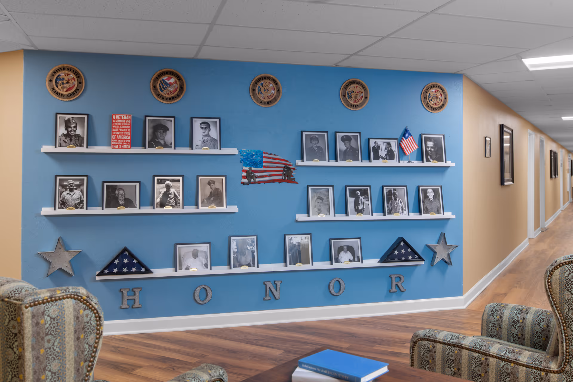 Interior common area with a blue 'HONOR' wall displaying framed veteran photos, flags, and plaques, with upholstered chairs in the foreground.