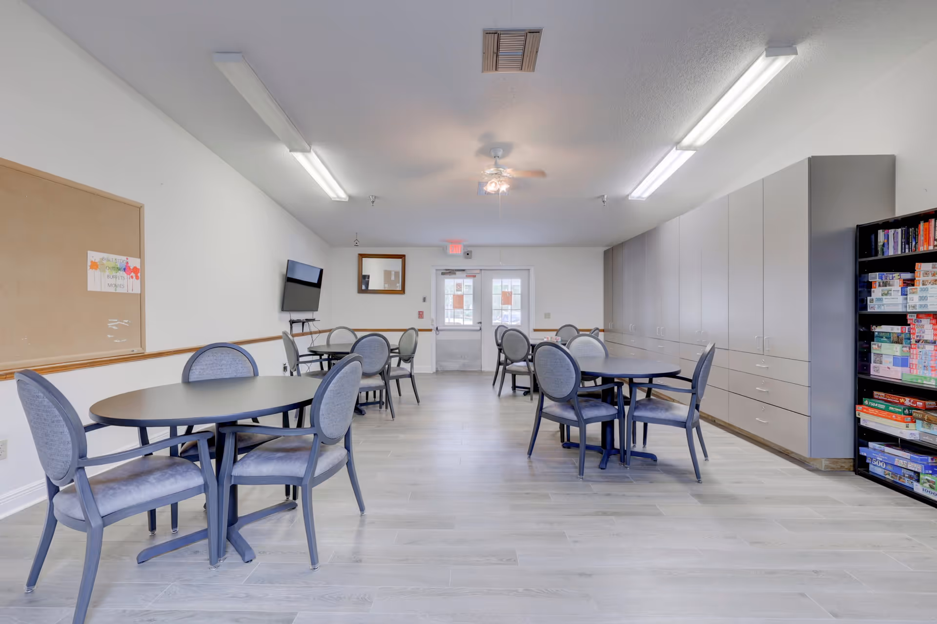 A bright and clean common room with several round tables and cushioned chairs arranged for group seating. The room features light-colored wood flooring, white walls, a bulletin board, a wall-mounted TV, ceiling lights, and a ceiling fan. On the right side, there are large cabinets and a bookshelf filled with board games.