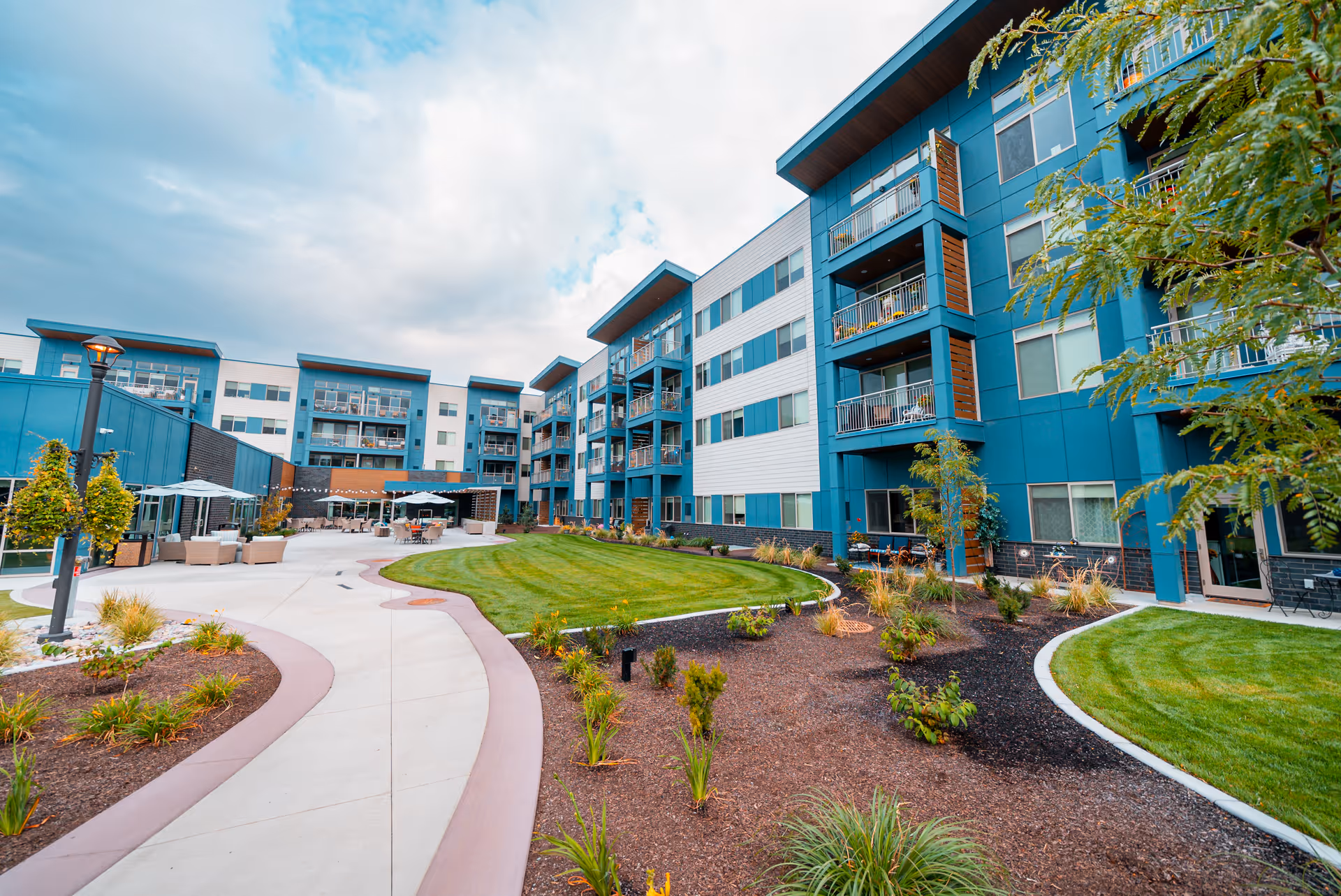 Outdoor courtyard area of a senior living facility with a curved concrete walkway, landscaped garden beds, green grass, and a multi-story building with balconies in the background under a partly cloudy sky.