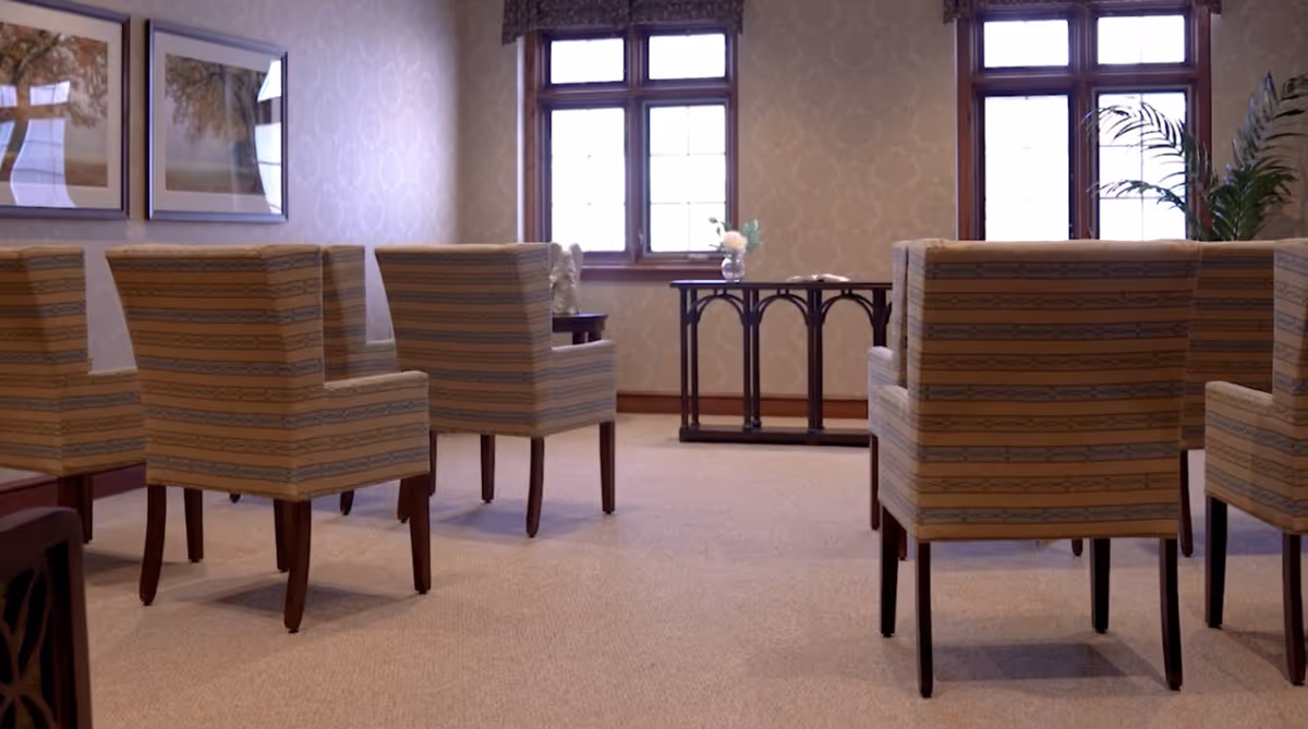 Rows of upholstered striped chairs arranged in a carpeted sitting room with windows and a small table at the front.
