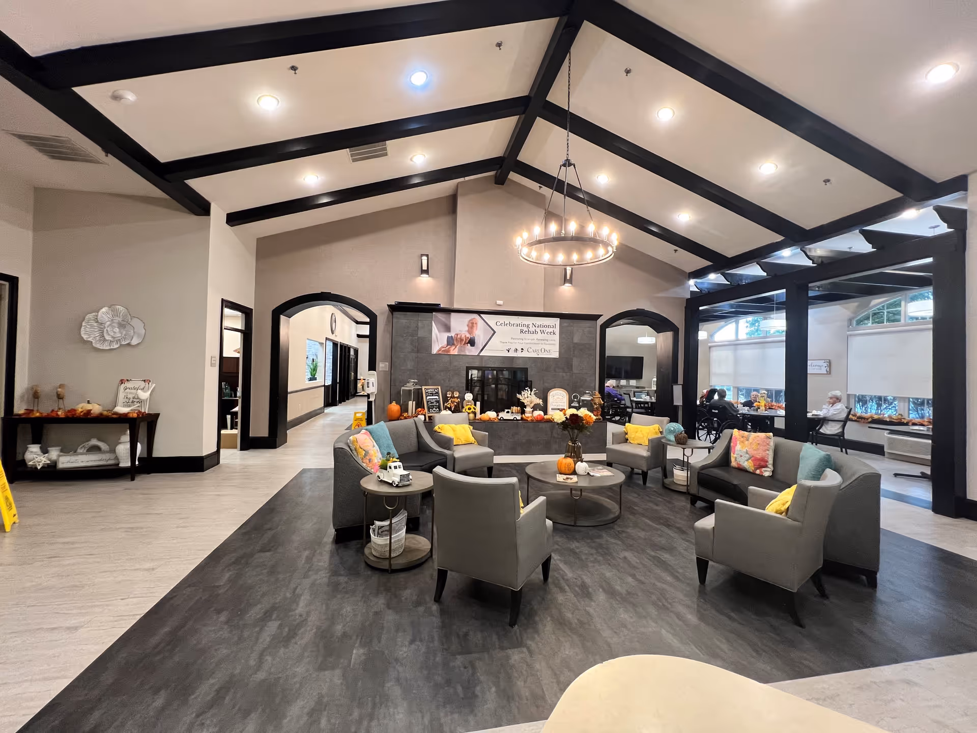 A spacious and well-lit assisted living facility common area with high vaulted ceilings featuring dark wooden beams. The room has a modern chandelier and recessed lighting. There are several comfortable chairs and sofas arranged around circular coffee tables decorated with pumpkins and flowers. In the background, a banner reads 'Celebrating National Rehab Week'. Large windows with blinds allow natural light into the space, where a few elderly residents are seated. The floor has a combination of light and dark wood-like finishes, and the walls are painted in neutral tones with black trim around doorways and windows.