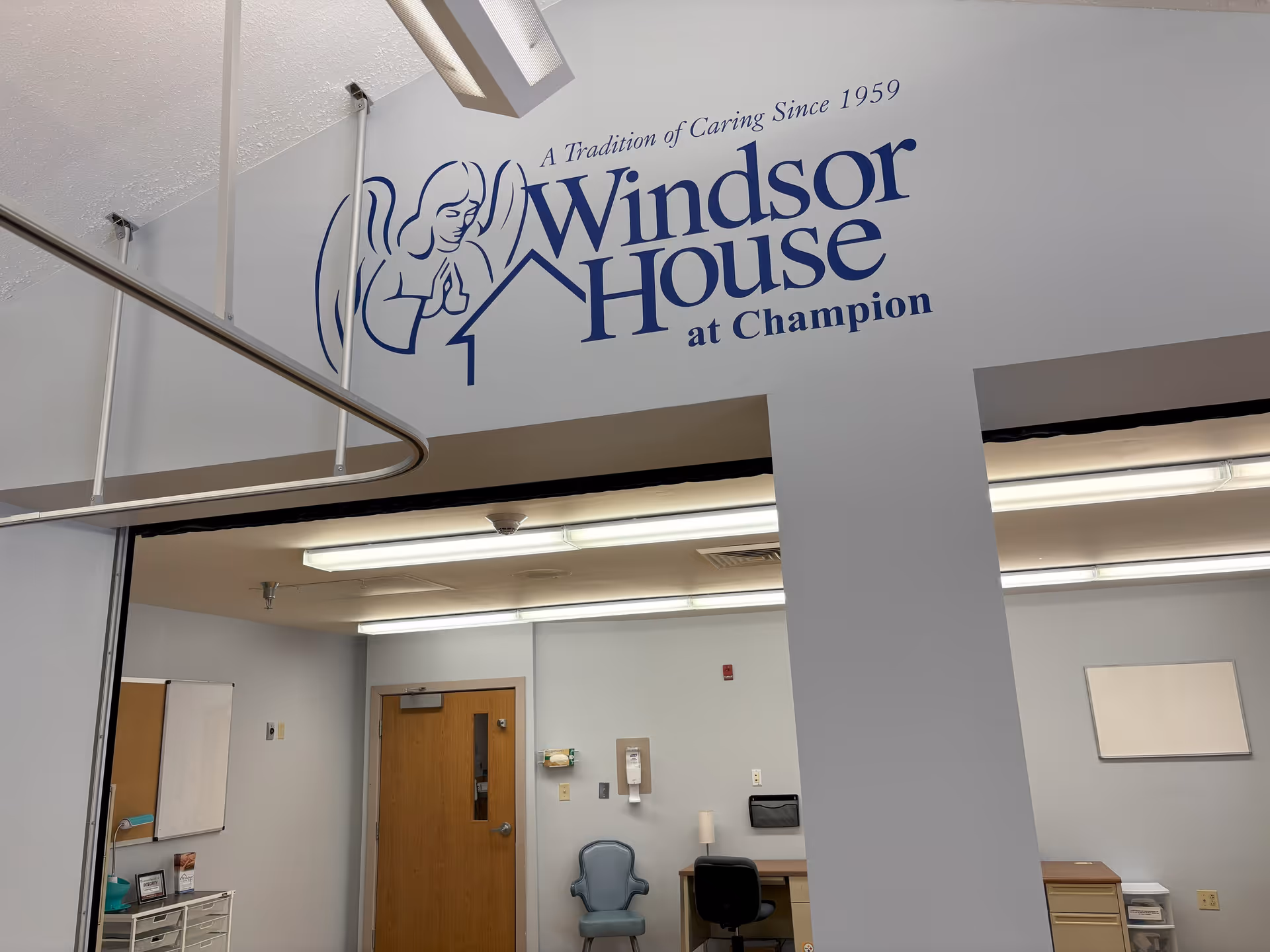 Interior view of a room at Windsor House at Champion showing a wall with the facility's logo and slogan 'A Tradition of Caring Since 1959'. The room has fluorescent ceiling lights, a wooden door, a blue chair, a desk with a black office chair, and some office supplies on the walls and surfaces.