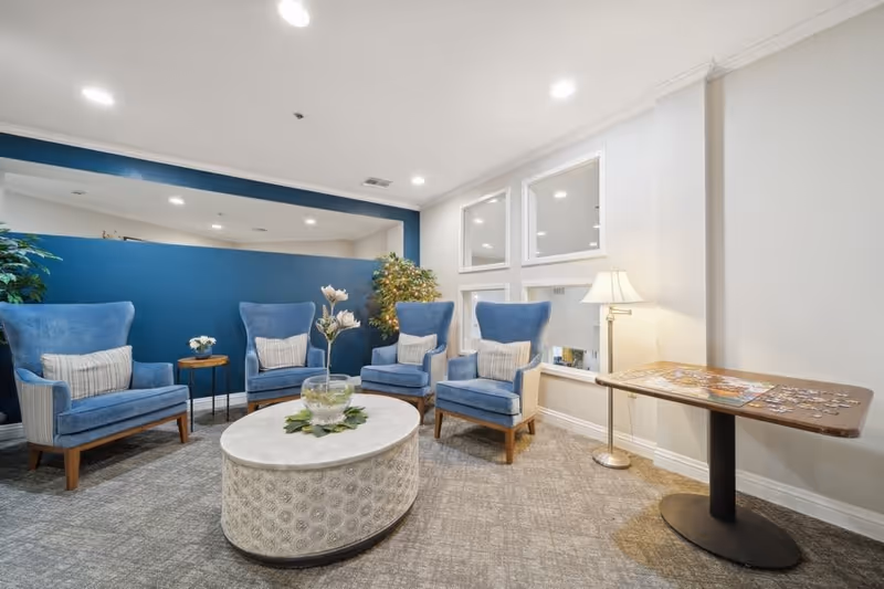 Bright common-area lounge with four blue wingback chairs arranged around a round coffee table and a lamp beside a puzzle-covered side table.