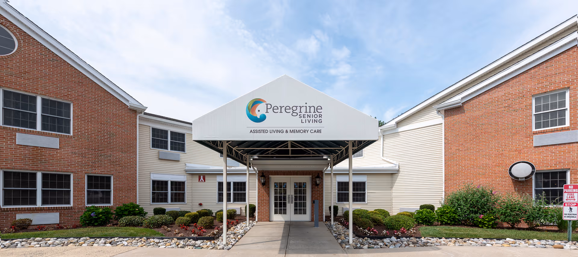 Front exterior view of Peregrine Senior Living at Salisbury building with a covered entrance, brick and siding walls, windows, and landscaped bushes and flowers under a partly cloudy sky.