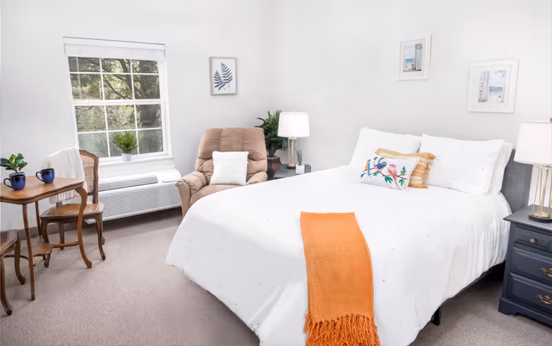 A bright and cozy bedroom featuring a large bed with white bedding and decorative pillows, including one with a bird design. An orange throw blanket is draped across the foot of the bed. To the right of the bed is a dark blue nightstand with a lamp. A comfortable beige armchair with a white pillow is positioned near a window with white blinds. A small wooden table with two blue mugs and a plant sits next to a wooden chair. The walls are white with framed artwork, and there is a green plant in the corner.