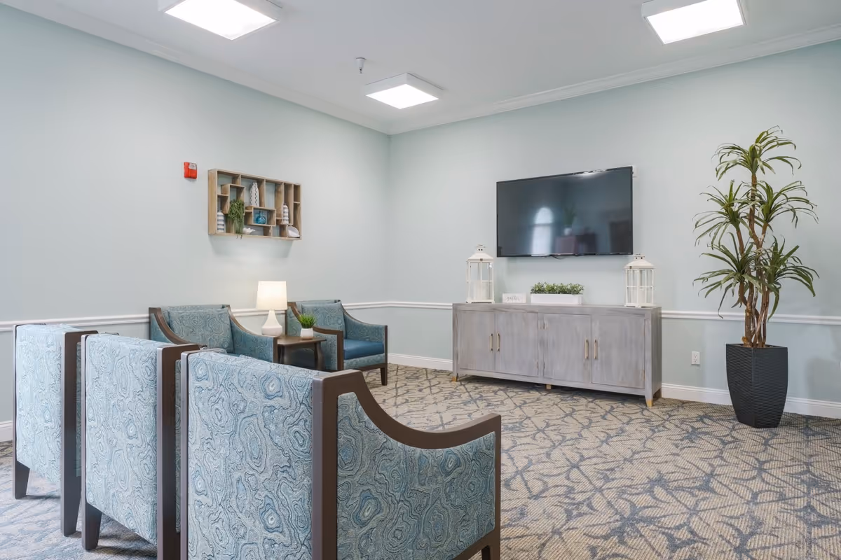 A cozy living room area with four patterned blue armchairs arranged facing a wall-mounted flat-screen TV. Below the TV is a light gray wooden cabinet decorated with two white lanterns and a small plant. A tall potted plant stands in the corner, and a small side table with a lamp and a small plant is placed between two of the chairs. The walls are painted light blue with white trim, and the carpet has a blue and beige geometric pattern.