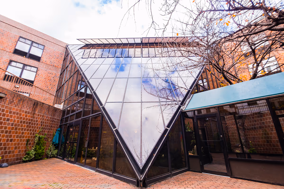 Exterior view of a building with a unique triangular glass facade reflecting the sky, surrounded by brick walls and windows, with some bare tree branches visible.