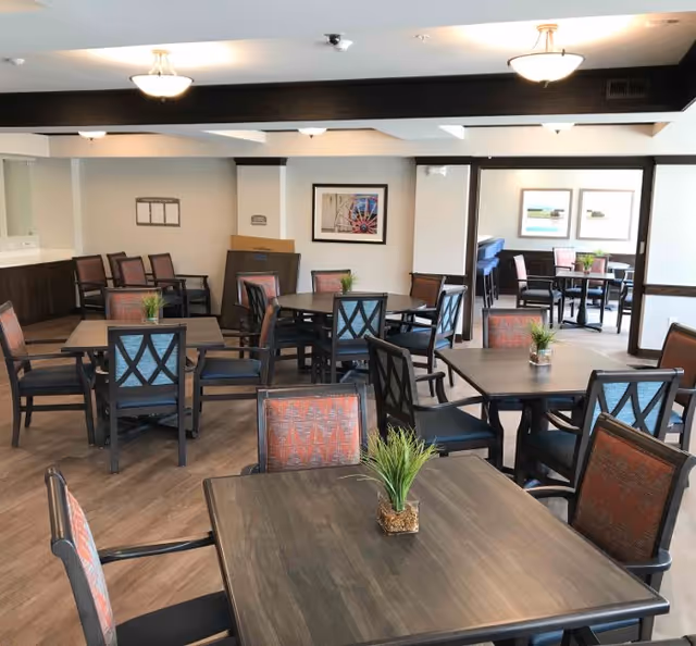 A dining room with multiple square wooden tables and chairs with patterned upholstery. Each table has a small green plant centerpiece. The room has light-colored walls, framed artwork, and ceiling lights providing illumination.