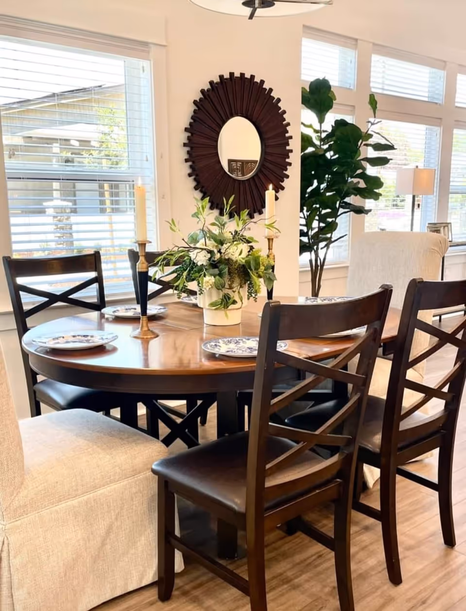 A dining area with a round wooden table set with four plates, two tall candles in holders, and a floral centerpiece. Surrounding the table are four dark wooden chairs with black cushions and one beige upholstered chair. In the background, there is a large window with white blinds, a decorative round mirror with a sunburst frame on the wall, a tall green plant, and a floor lamp.