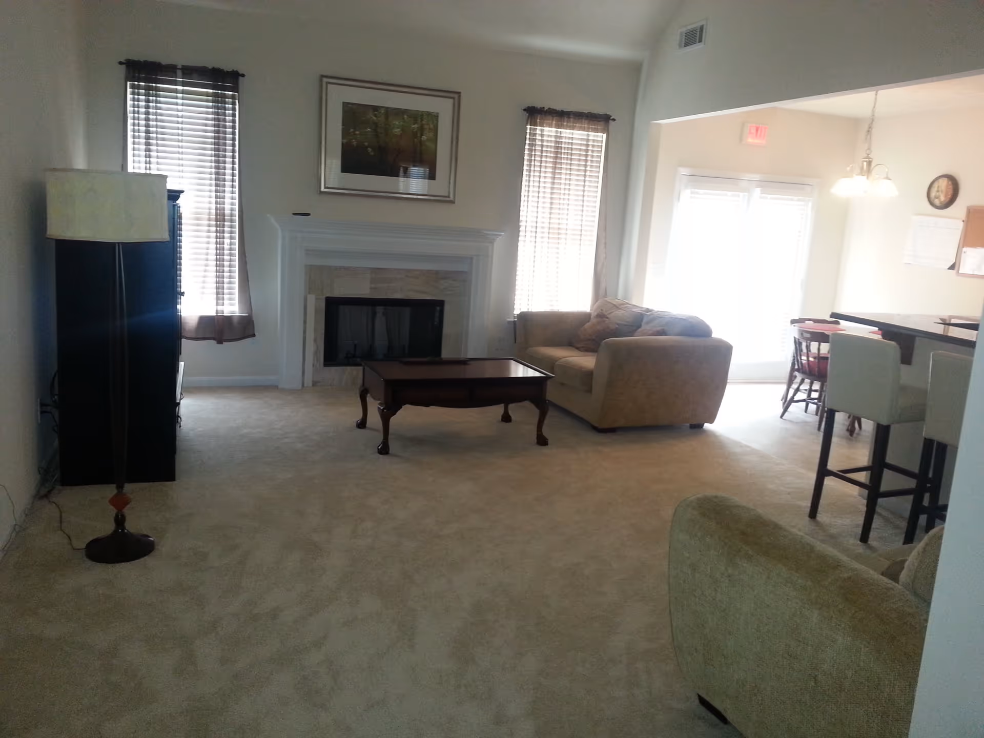 Carpeted living room with a fireplace, coffee table, sofa, floor lamp and an adjacent dining area with bar stools.
