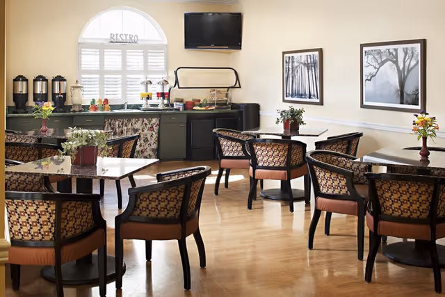 A cozy dining area with several tables and cushioned chairs arranged neatly. The room has light-colored walls adorned with framed black and white nature photographs. A counter at the back holds beverage dispensers, a small refrigerator, and a TV mounted above. A window with white shutters lets in natural light, and small flower arrangements decorate the tables.