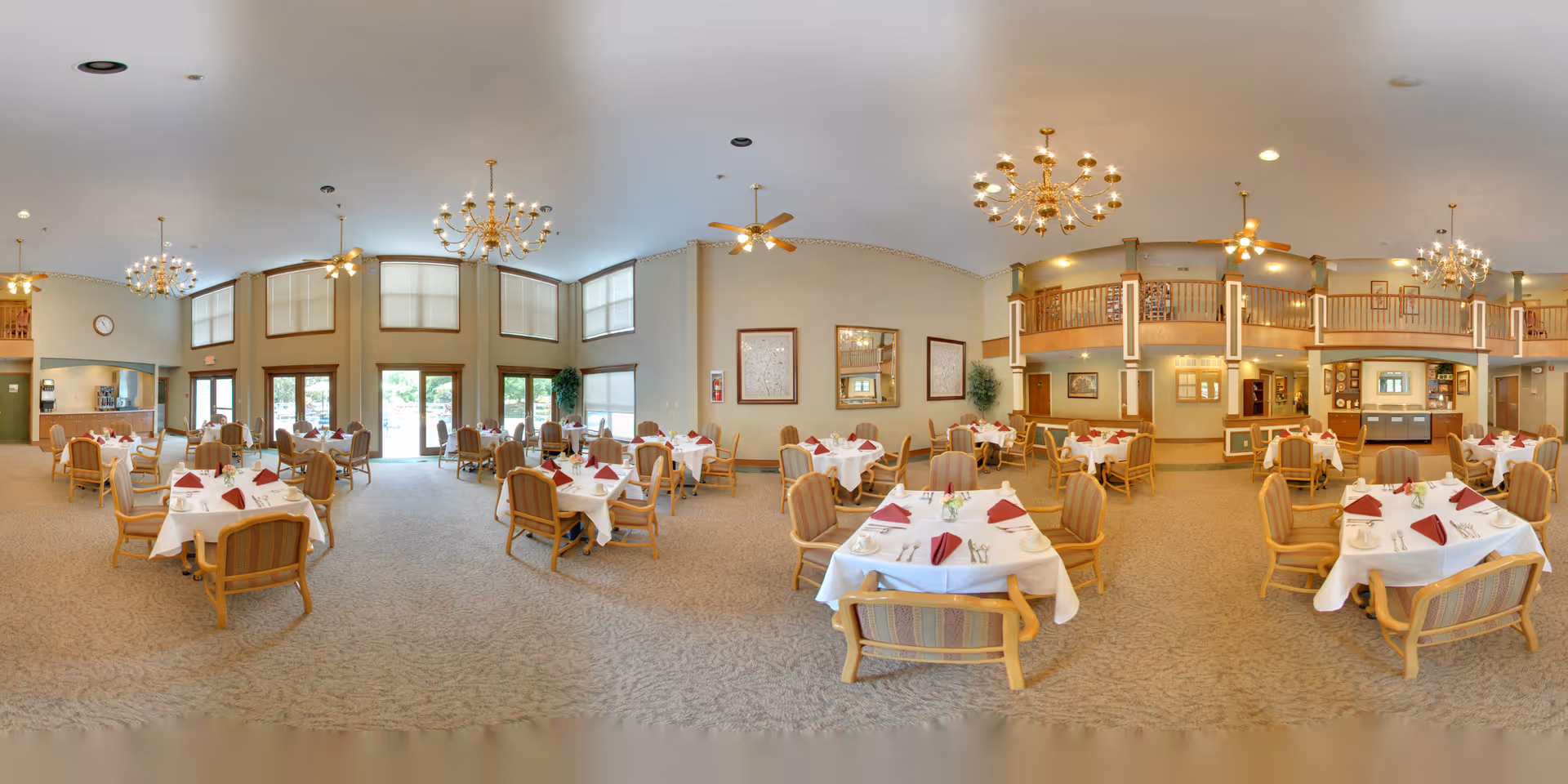 A spacious dining room in a senior living facility with multiple tables covered in white tablecloths and set with red napkins, silverware, and small flower arrangements. The room features high ceilings with chandeliers and ceiling fans, large windows allowing natural light, and a balcony overlooking the dining area. The decor includes framed artwork on the walls and neutral-toned carpeting.