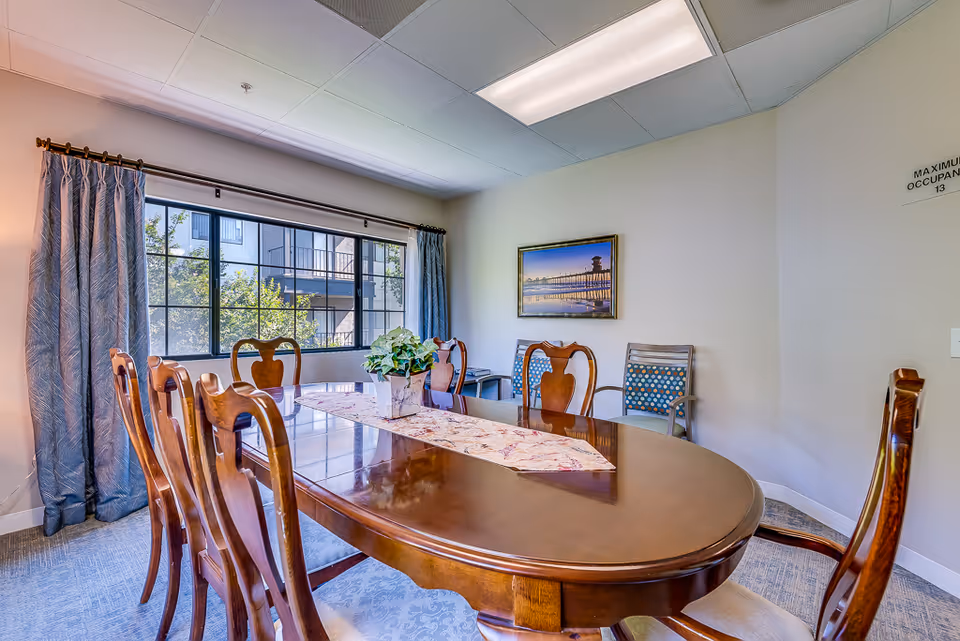 A bright dining room with a polished wooden table and chairs, a centerpiece plant, large window with curtains, and wall art.