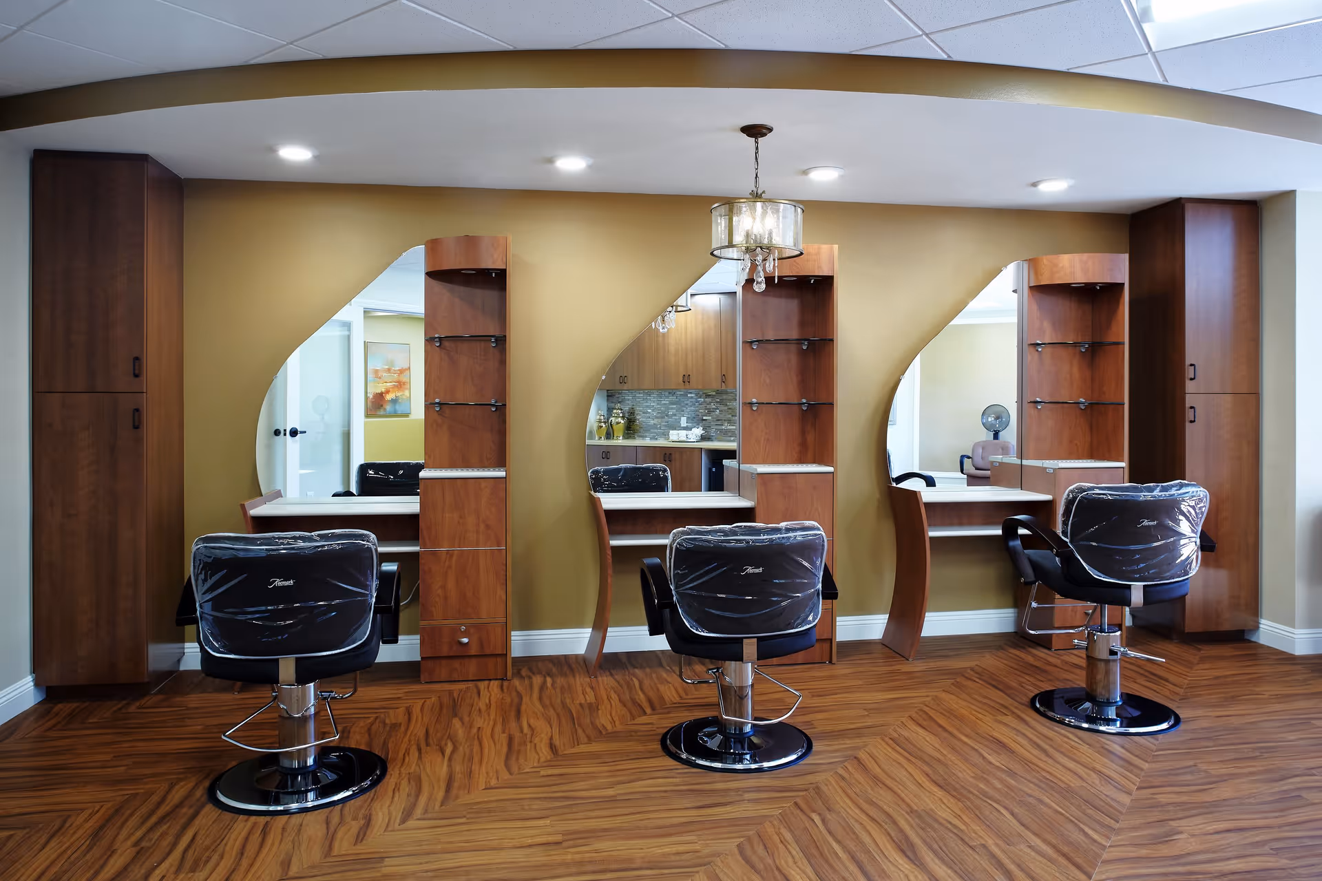 Interior view of a salon area with three styling stations, each featuring a large mirror, wooden shelving, and a black salon chair with protective plastic covering. The floor has a wood pattern, and the walls are painted a warm beige color. A chandelier-style light fixture hangs from the ceiling.