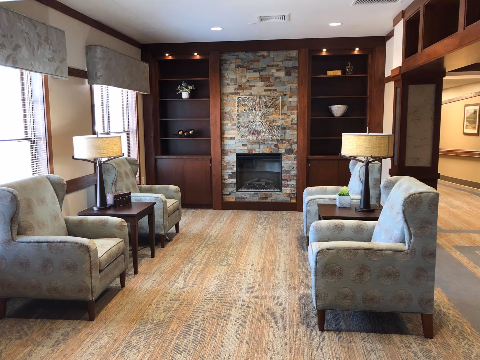 A cozy sitting area with four patterned armchairs arranged in pairs facing each other, separated by two wooden side tables with lamps. The room features large windows with blinds and valances on the left side, a stone fireplace with a decorative metal artwork above it, and built-in wooden shelves on either side of the fireplace. The floor is covered with a patterned carpet, and the walls have wooden trim and neutral paint.