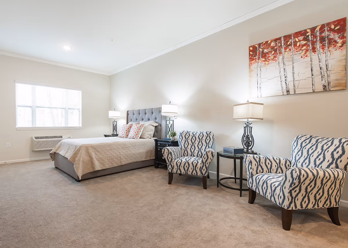 A bright and spacious bedroom with a large bed featuring a tufted headboard, two patterned armchairs, a small round side table with a lamp and books, and a large window letting in natural light. A colorful painting of trees with red leaves hangs on the wall above the chairs.