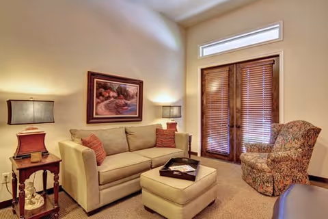 A cozy living room with a beige sofa adorned with two red patterned pillows, a matching ottoman with a tray on top, a floral-patterned armchair, two wooden side tables each with a lamp, a framed painting on the wall, and wooden blinds covering a glass door with a transom window above.