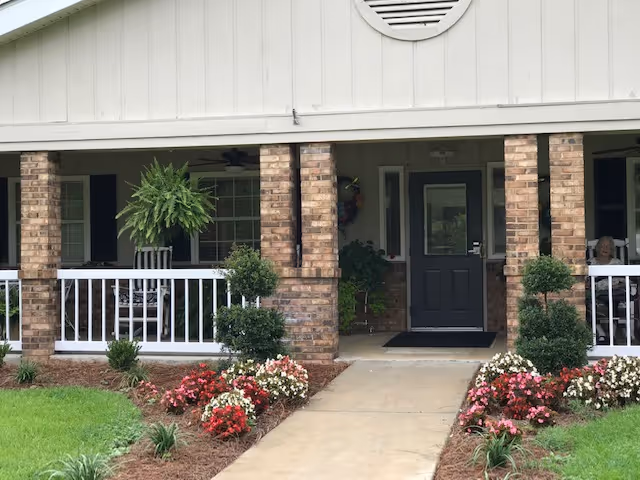Front porch entrance with brick columns, a black door, white railing, hanging fern, and flower beds lining a concrete walkway.