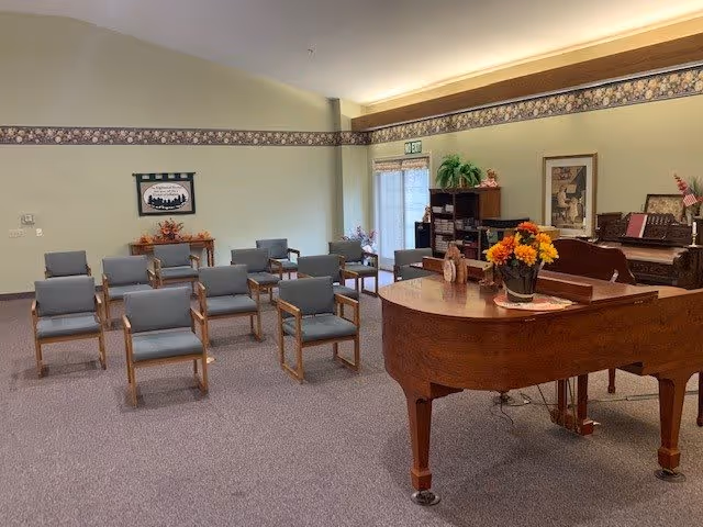A room with several rows of gray cushioned chairs with wooden armrests arranged facing a grand piano. The room has light green walls with a floral border near the ceiling, a framed picture on the wall, and a bookshelf with plants and decorations near a door with a window. There is also a wooden table with a flower arrangement on the piano.