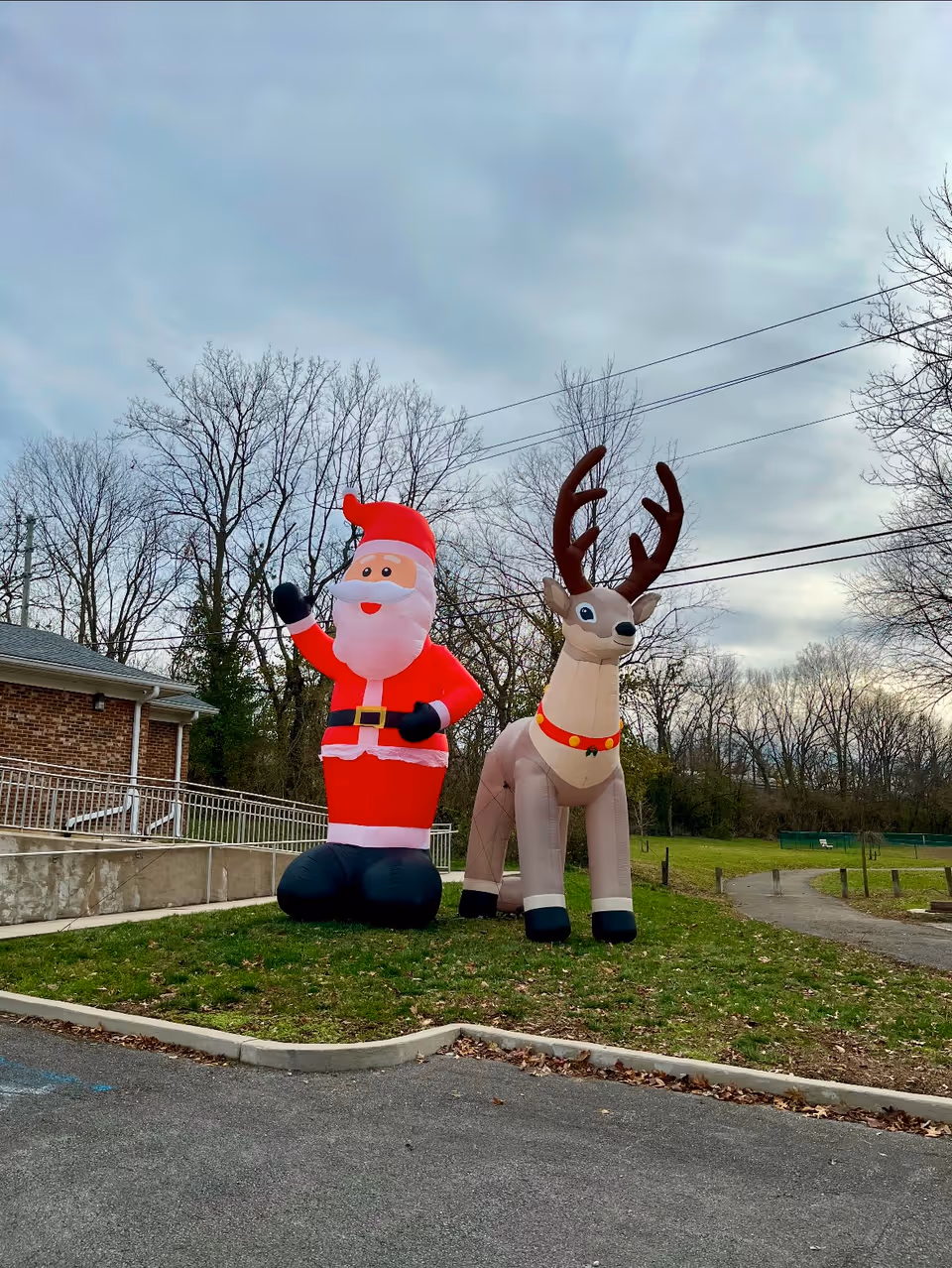 Outdoor scene showing large inflatable decorations of Santa Claus and a reindeer on a grassy area near a building with leafless trees and a cloudy sky in the background.