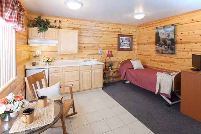 A cozy room with wooden paneled walls featuring a single bed with a maroon bedspread and a pillow, a small wooden nightstand with a lamp and framed picture, a wooden cabinet with a TV on top, a kitchenette with light wood cabinets and a sink, and a small round table with two mugs and a flower arrangement next to a wooden chair with a cushion. The floor is partly tiled and partly carpeted, and there are two ceiling lights providing illumination.