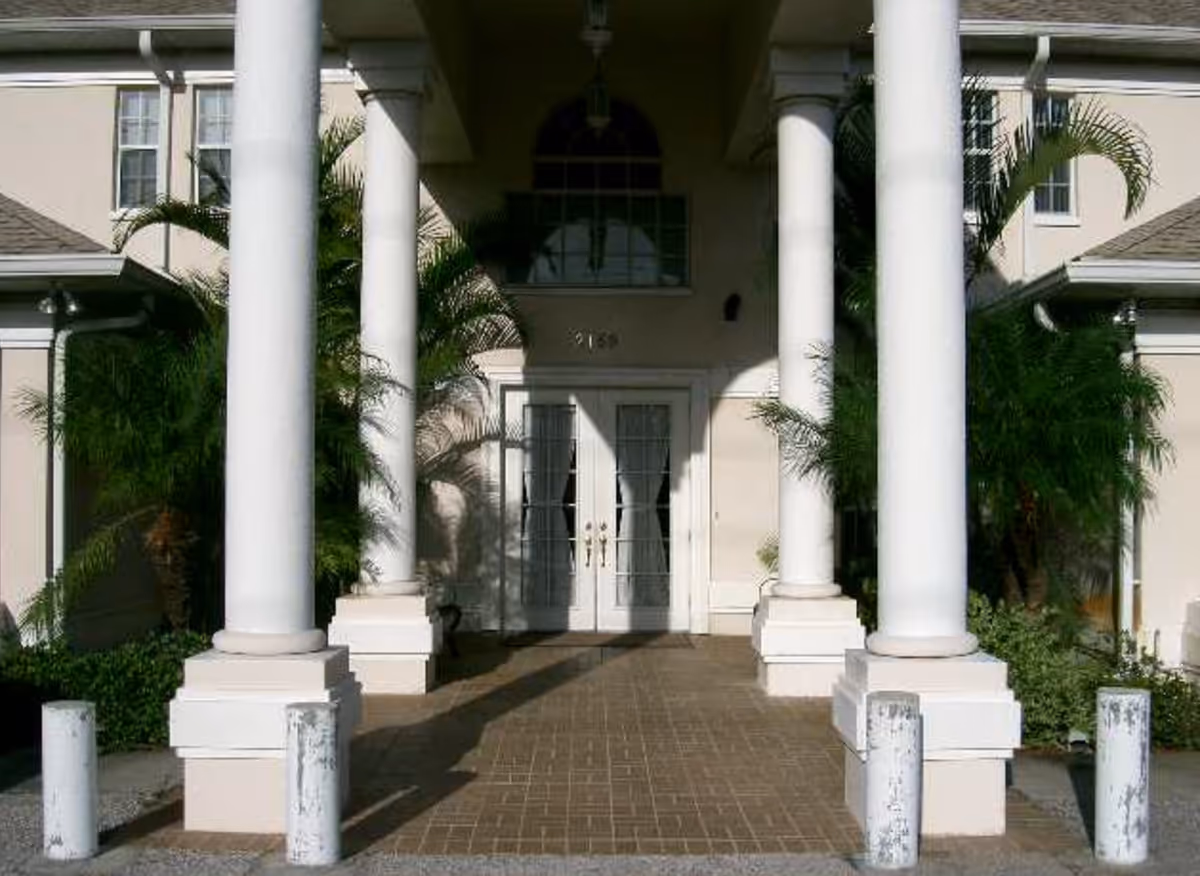 Entrance of a building with four large white columns, double glass doors, and surrounding greenery including palm plants.