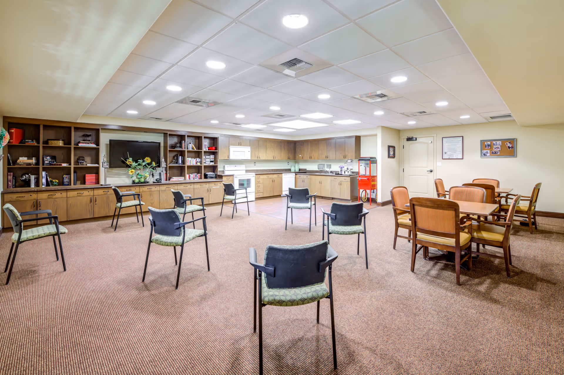 A spacious common room with a kitchen area along the back wall featuring wooden cabinets, a stove, microwave, and dishwasher. Several chairs are arranged in a circle in the center of the room, and there are two tables with chairs on the right side. The room is well-lit with ceiling lights and has a carpeted floor.