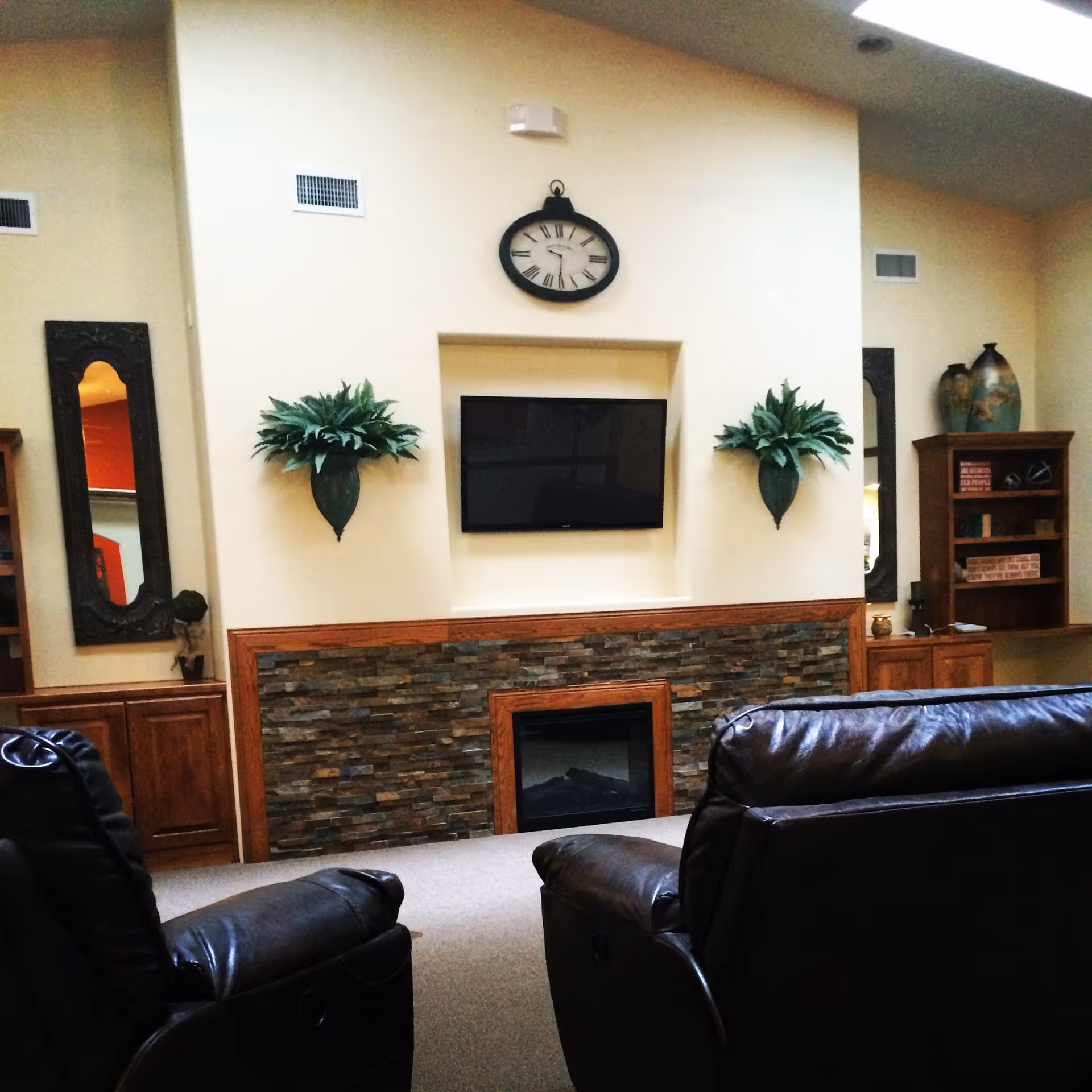 A cozy living room area with two dark leather recliners facing a wall-mounted flat screen TV above a stone fireplace. The wall is decorated with two green plants in wall-mounted planters and a large clock above the TV. Wooden cabinets and shelves with decorative items flank the fireplace on both sides.