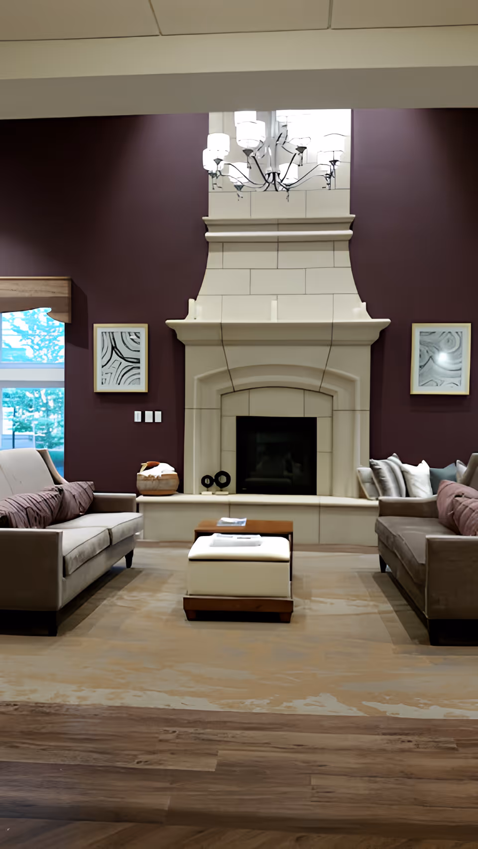 A cozy living room with two beige sofas facing each other, a white cushioned ottoman in the center, and a large white stone fireplace against a dark purple wall. Above the fireplace hangs a modern chandelier, and there are two abstract framed artworks on either side of the fireplace. A window with a wooden valance is visible on the left side.