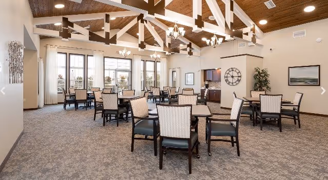 A spacious dining room in a senior living facility with multiple tables and chairs arranged neatly. The room features a high ceiling with exposed wooden beams, large windows allowing natural light, chandeliers, a wall clock, and framed artwork on the walls.