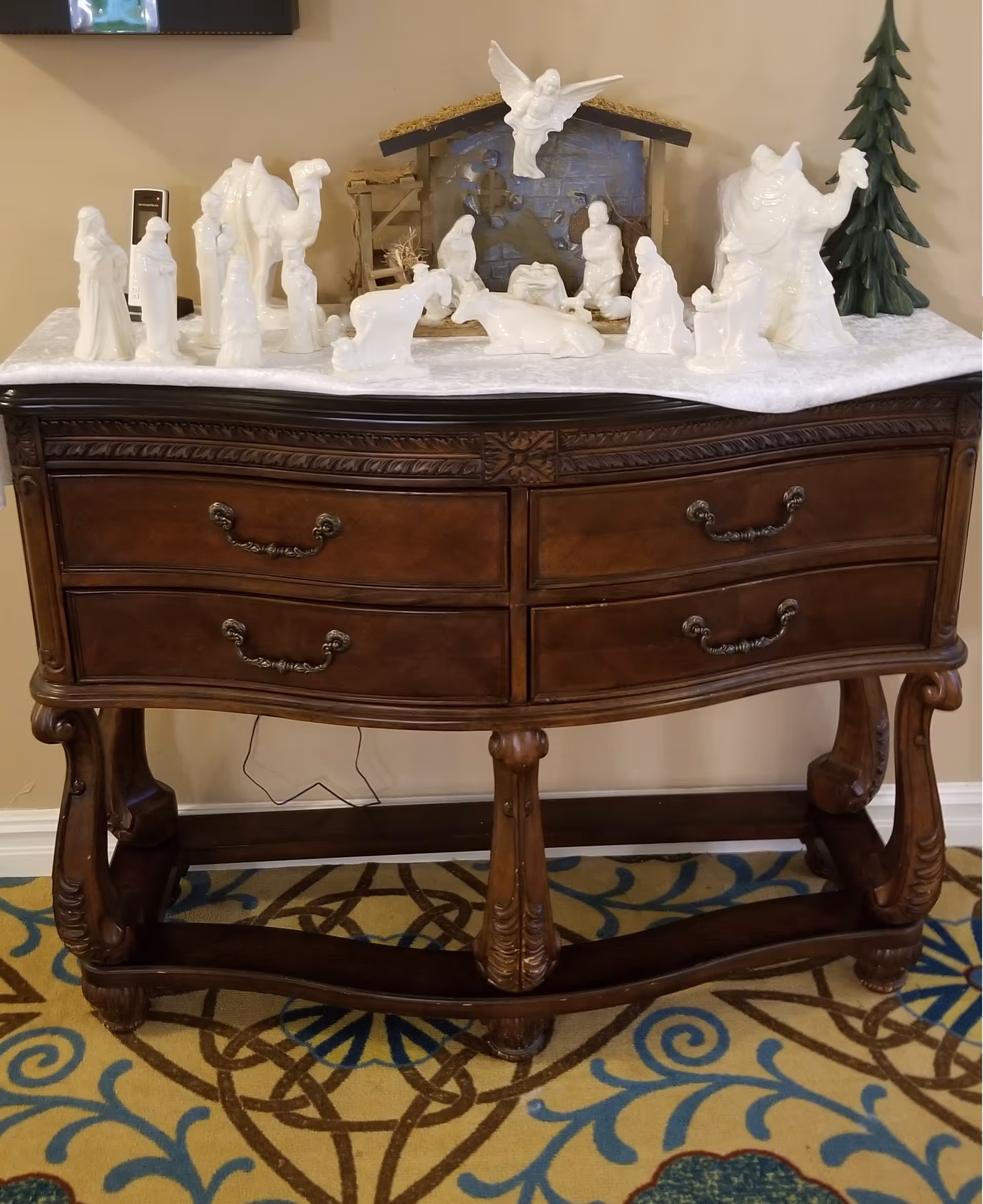 Ornate wooden console table topped with a white ceramic nativity scene and a small stable against an interior wall.