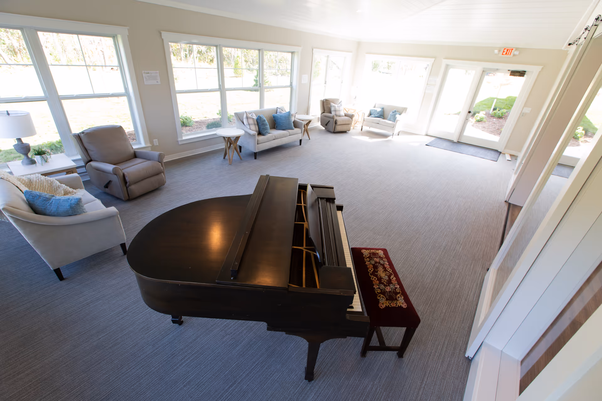 Bright communal living room with a grand piano, cushioned sofas and recliners, and large windows and doors.