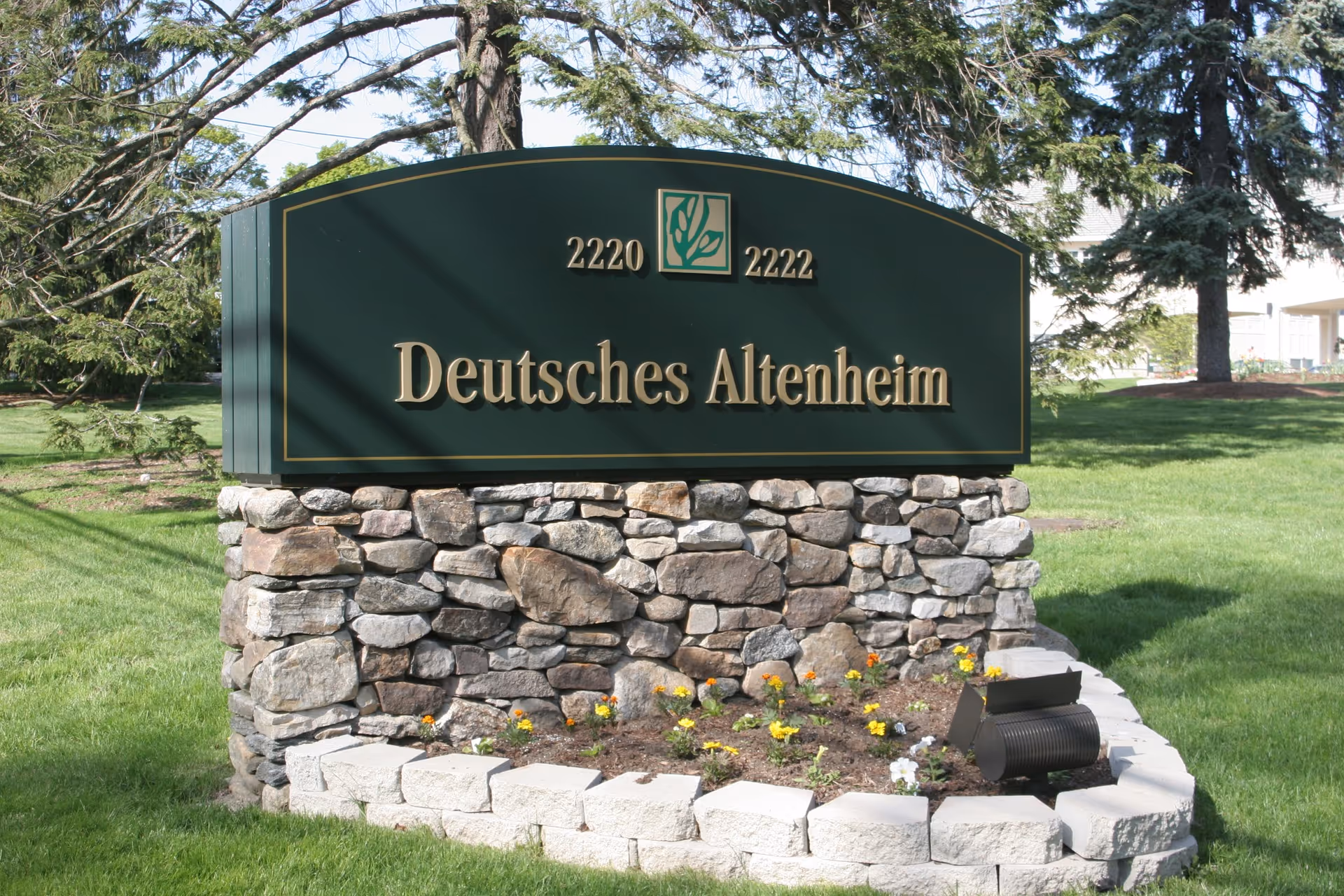 A stone and brick sign on a grassy area with trees in the background. The sign reads 'Deutsches Altenheim' with the numbers 2220 and 2222 on either side of a green and white logo.