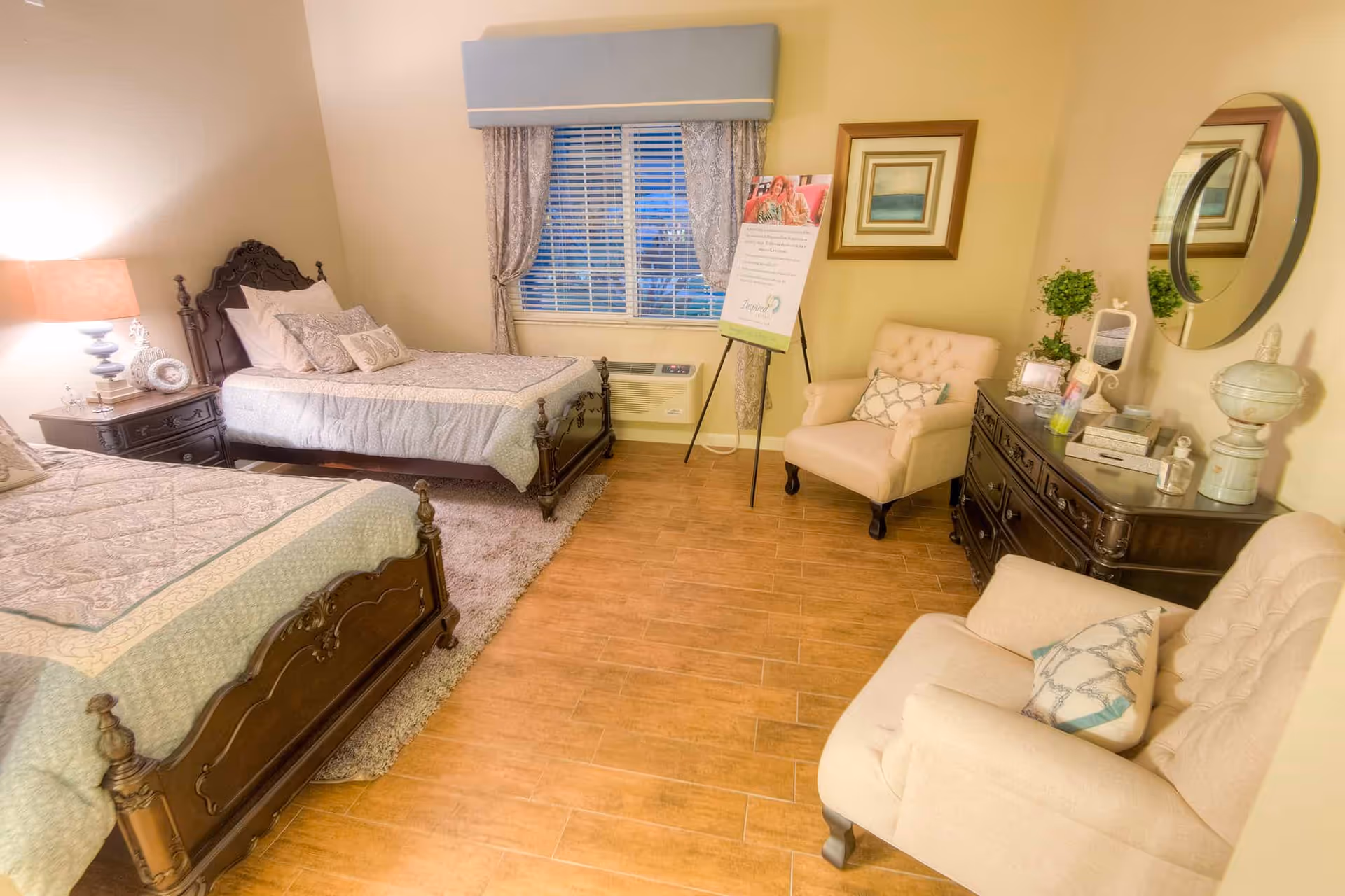 A cozy bedroom with two wooden twin beds covered in light-colored bedding. There is a nightstand with a lamp between the beds. The room has a window with curtains and a blue valance. Two beige armchairs with decorative pillows are placed near a dark wooden dresser with a round mirror above it. A framed picture hangs on the wall, and a standing sign is positioned near the window.