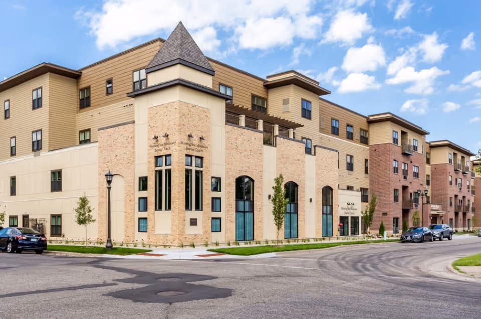 Exterior street-level view of a multi-story beige-and-brick senior living building with large windows and parked cars.