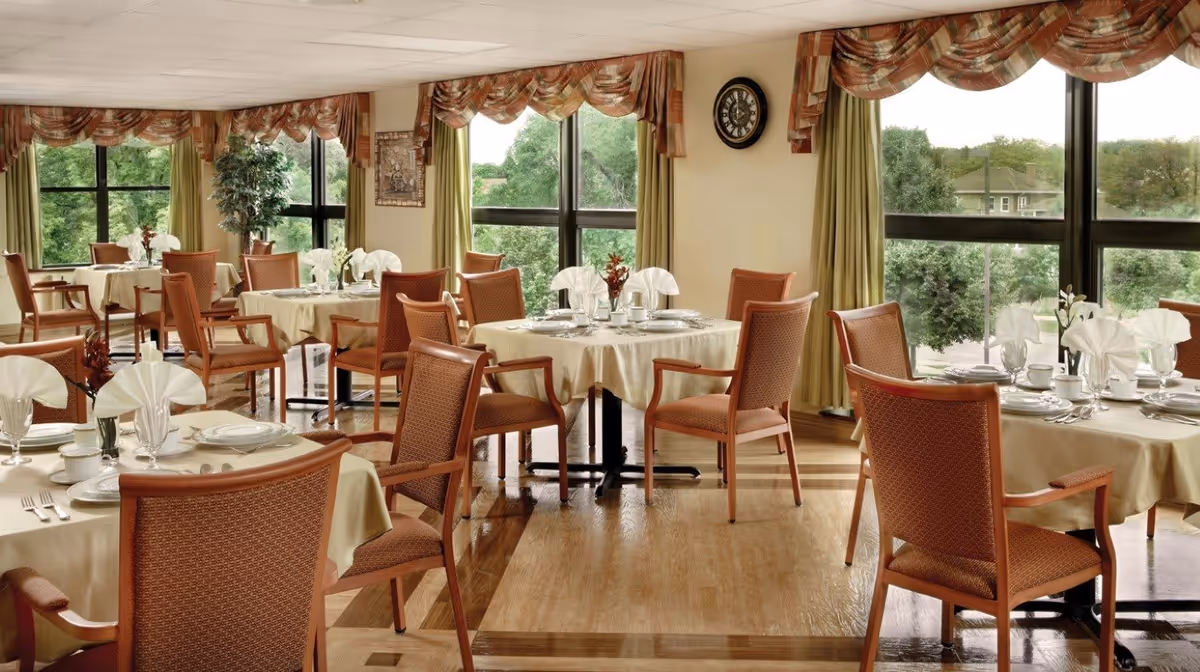 A bright dining room with several round tables covered with beige tablecloths, each set with white plates, cups, silverware, and folded white napkins. The room has large windows with green curtains and patterned valances, allowing natural light to fill the space. Wooden chairs with orange upholstery surround the tables, and a clock is mounted on the wall.