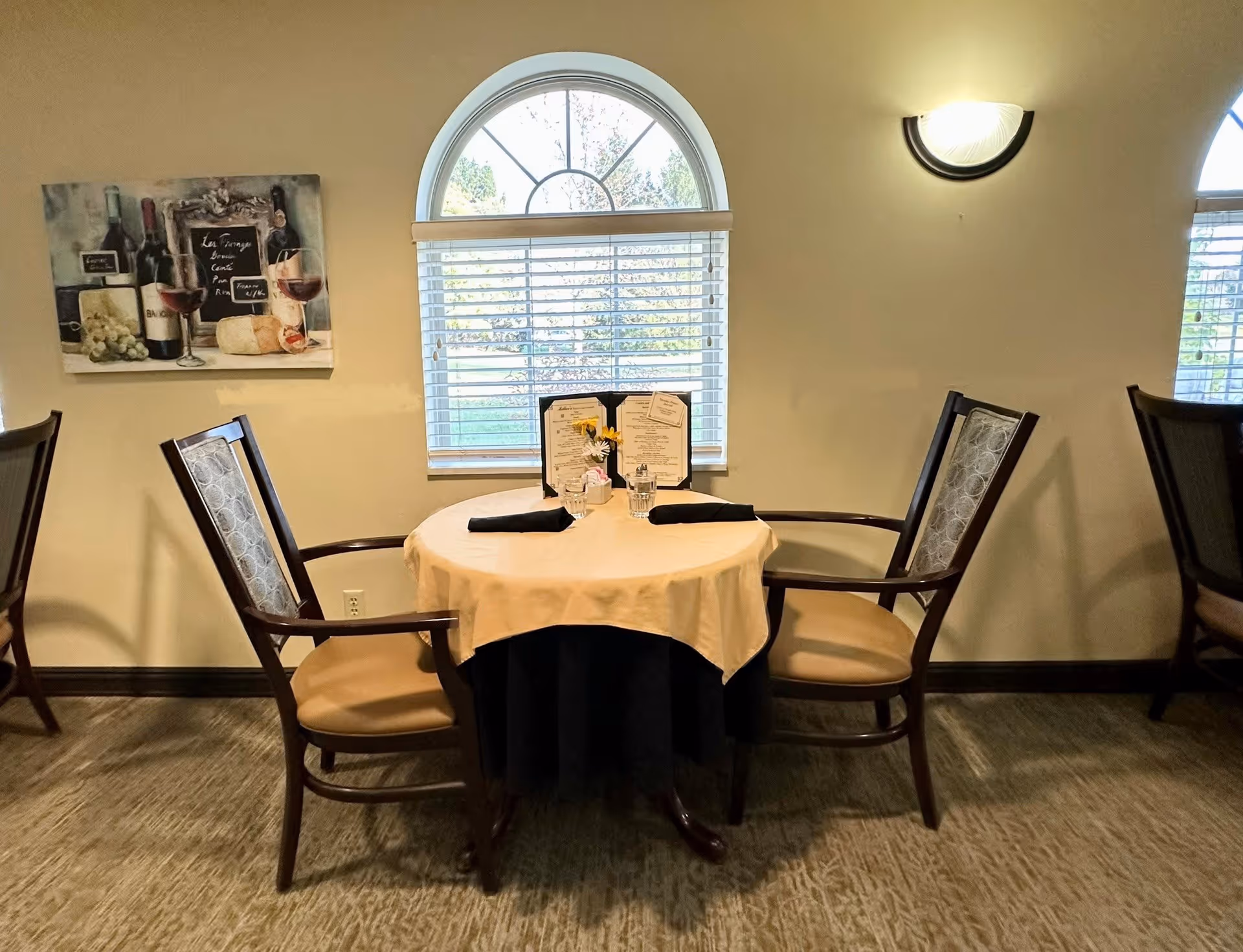 A small round dining table set for two with a beige tablecloth and black napkins, placed in front of a window with white blinds and an arched top. Two wooden chairs with cushioned seats and patterned backs are positioned on either side of the table. A wall-mounted light fixture is above the table, and a painting of wine bottles, glasses, and cheese hangs on the wall to the left.