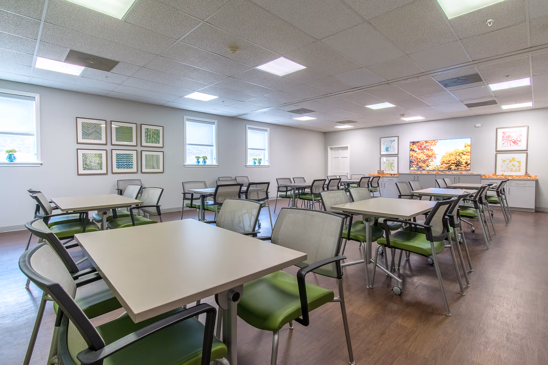 A bright and clean dining room with multiple square tables and green cushioned chairs arranged neatly. The room has white walls adorned with framed artwork and three windows letting in natural light. A large TV screen on the far wall displays an autumn scene, and the floor is a dark wood finish.